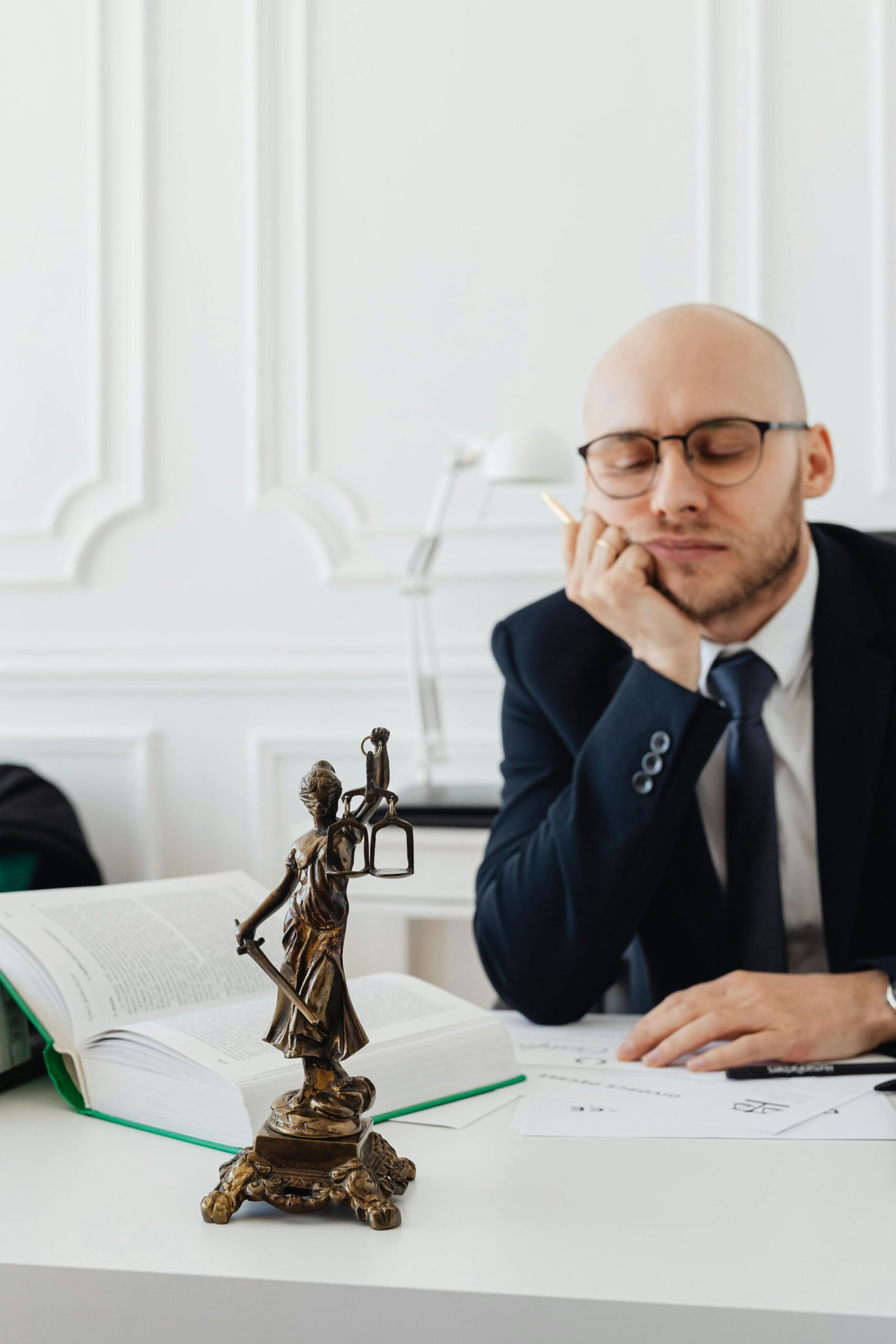 Home A thoughtful lawyer sits at his desk beside a statue of Lady Justice, conveying contemplation.